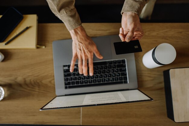 Ręka mężczyzny trzyma kartę kredytową i korzysta z laptopa w domu, pracujący biznesmen lub przedsiębiorca, online