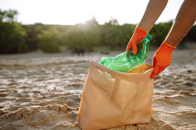 Ręka mężczyzny zbiera plastikową butelkę na plaży. Wolontariusz w rękawicach ochronnych zbiera plastik butelki.