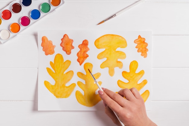 Ręka rysująca jesienne liście różne kształty, rozmiary i kolory z akwarelami i pędzlem na białym stole Widok z góry