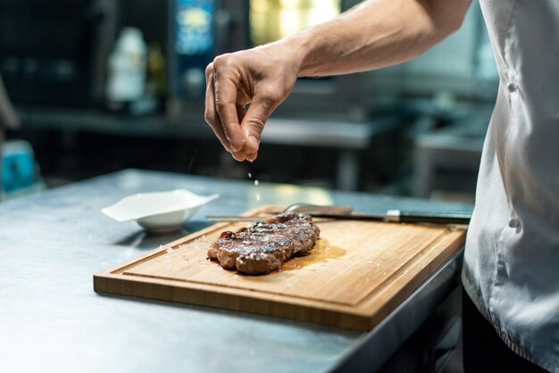 Ręka szefa kuchni posypuje przyprawy na pieczonym steku wołowym na drewnianej desce, stojąc przy stole i przygotowując posiłek dla klienta restauracji