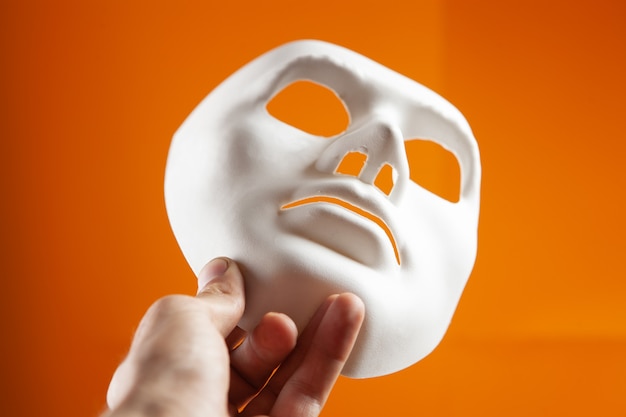 ręka trzyma białą anonimową maskę na pomarańczowym tle
