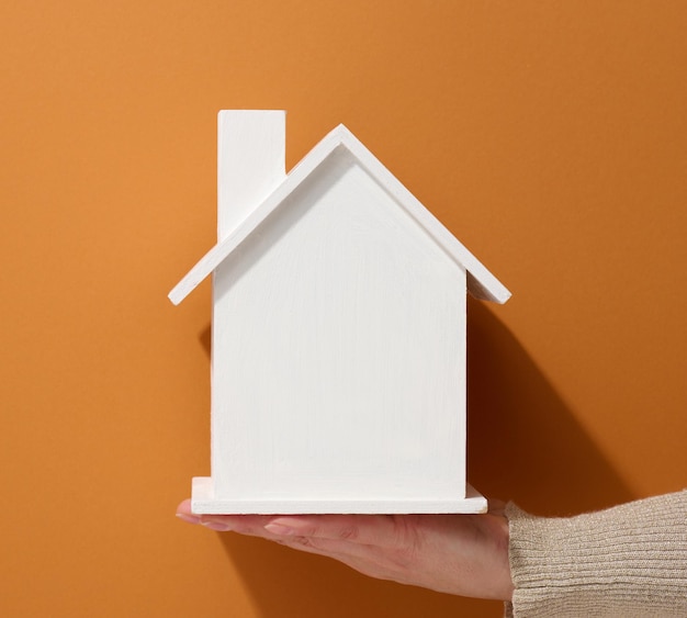 Ręka trzyma biały miniaturowy drewniany dom na brązowym tle. Koncepcja ubezpieczenia nieruchomości, ochrona środowiska, szczęście rodzinne