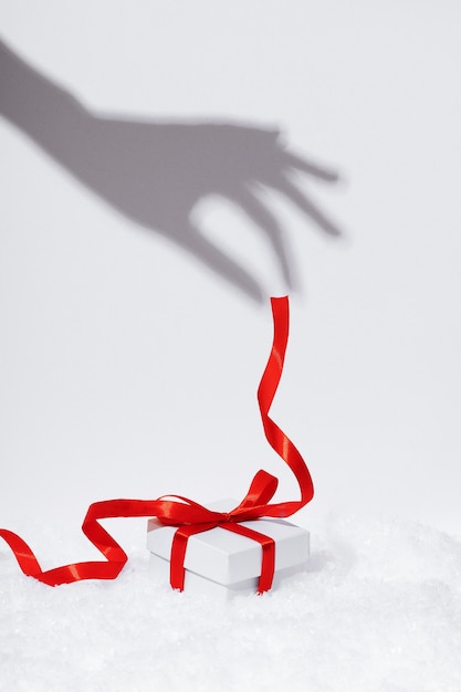 Zdjęcie ręka trzyma cień czerwoną wstążką prezent na białym tle. białe pudełko do pakowania prezentów na śnieżnym tle. świąteczna kompozycja z wystrojem na nowy rok.