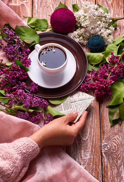 Ręka trzyma filiżankę porannej kawy z gałęzi wiosennych kwiatów bzu kwitnących na drewniane tła widok z góry.