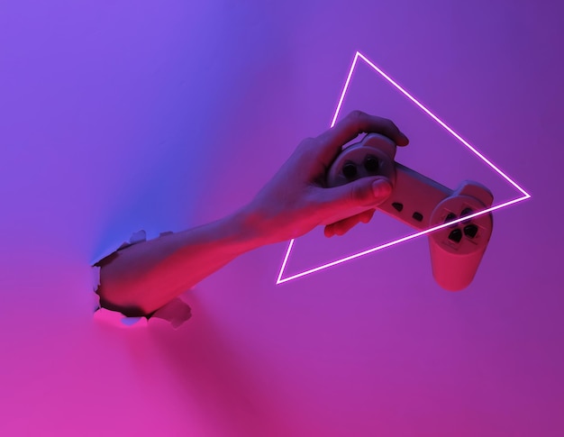 Ręka trzyma gamepada przez rozdarty otwór. Kreatywny pop-art różowy niebieski neonowy kolor. Modne oświetlenie gradientowe. Nocne światło