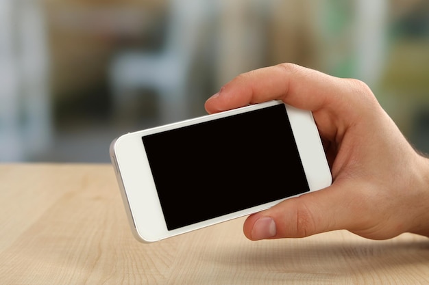 Ręka trzyma inteligentny telefon komórkowy na drewnianym stole i jasnym niewyraźnym tle