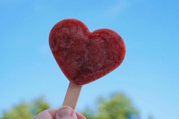 Ręka trzyma popsicle w kształcie serca lub lody na patyku przed jasnym, błękitnym niebem