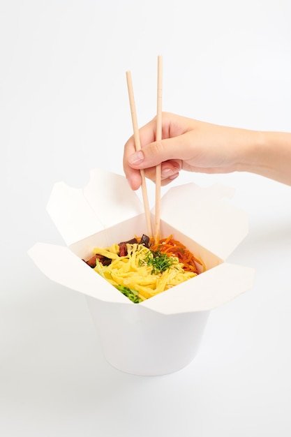 Ręka trzyma ryżowy wok z drewnianymi pałeczkami, azjatycki chiński wok na białym tle. Jedno otwarte pudełko na wynos z wokiem, koktajlem z owoców morza i warzywami. Koncepcja szybkiej dostawy żywności