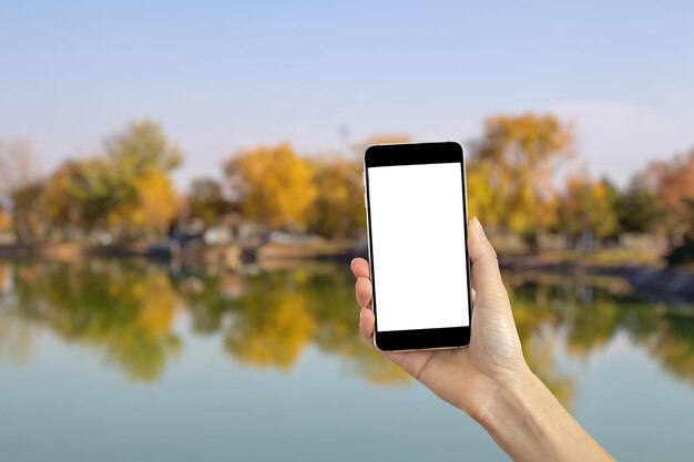 Ręka trzyma smartfona z pustym białym ekranem. Rozmazany widok na jezioro w tle. koncepcja podróży.