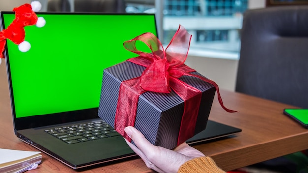 Ręka trzymająca duże pudełko z prezentami obok laptopa z zielonym ekranem makiety zakupów świątecznych
