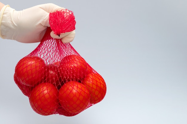 Ręka w gumowych rękawiczkach trzyma siatkę ze świeżymi pomidorami na szaro