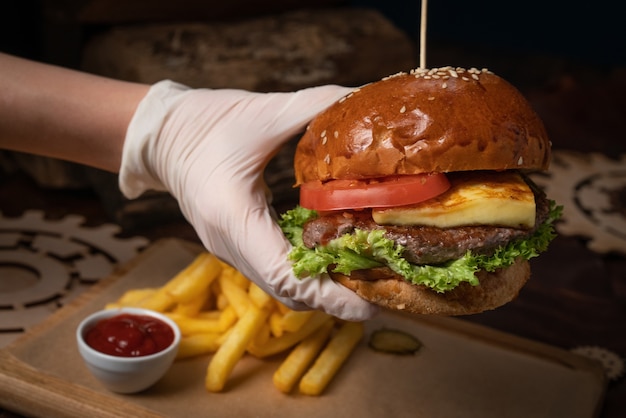 Ręka w jednorazowym blasku trzymająca świeżego rzemieślniczego burgera wołowego ze smażonym serem, podawana ze świeżą sałatą, frytkami i sosem pomidorowym.