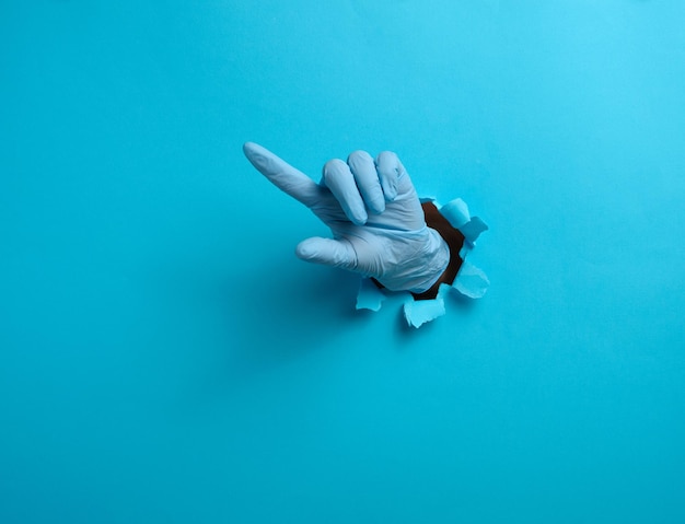 Zdjęcie ręka w niebieskiej rękawiczce medycznej wystaje z rozerwanej dziury na niebieskim tle papierowym