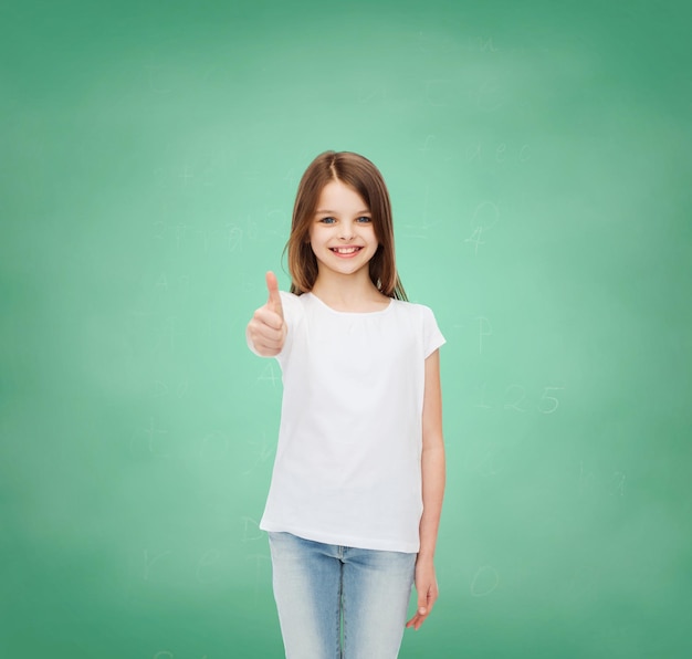 reklama, gest, edukacja, dzieciństwo i ludzie - uśmiechnięta dziewczynka w białej koszulce pokazująca kciuk w górę na zielonym tle tablicy