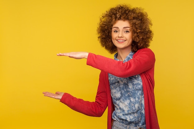 Reklama produktu Szczęśliwa urocza kobieta z kręconymi włosami prezentująca miejsce na kopię między rękami trzymająca puste miejsce na tekst reklamowy lub obraz w pomieszczeniu studio strzał na białym tle na żółtym tle
