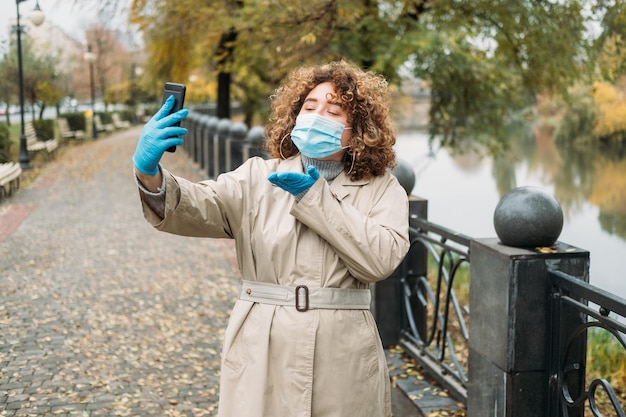 Relacja na odległość Blokada Covid Granice zamknięte Komunikacja mobilna Szczęśliwa kobieta z nadwagą w medycznej masce na twarz, która ma wideorozmowę wysyłającą buziaki w jesiennym parku