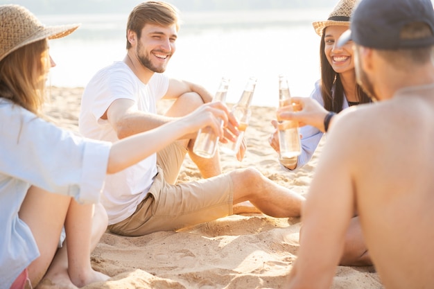 Relaks z najlepszymi przyjaciółmi. Grupa szczęśliwych młodych ludzi siedzących razem na plaży, rozmawiających i pijących piwo