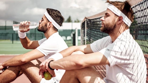 Relaksujący się po dobrej grze wesoły panowie przy siatce do tenisa i picie wody, podczas gdy jedno i drugie