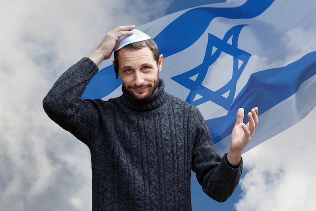 Zdjęcie religijny żyd z uśmiechniętą twarzą trzymający kippę na głowie na tle izraelskiej flagi piękne niebo przystojny uśmiechnięty brodaty żyd trzymający jarmulkę dla wiatru nad głową