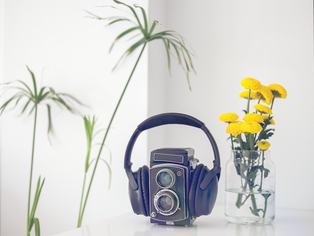 Retro czarny aparat fotograficzny ze słuchawkami umieszczonymi na białym stole z żółtymi kwiatami w szklanym wazonie w jasnym pokoju w domu