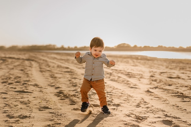 Roczny chłopiec uczy się chodzić po piasku nad rzeką.