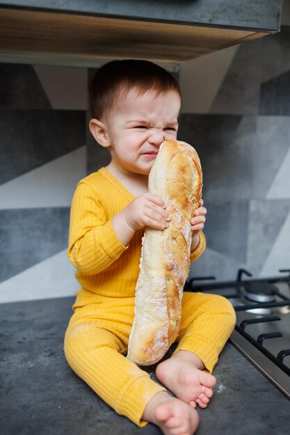 Roczny mały chłopiec w żółtych ubraniach siedzi i je świeżo upieczony chleb żytni Dziecko trzyma w rękach świeżą bagietkę