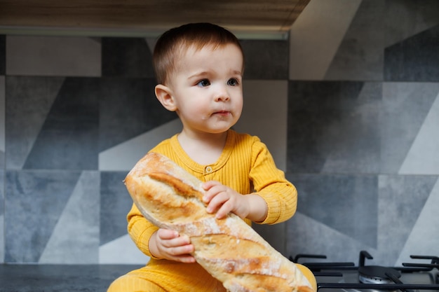 Roczny mały chłopiec w żółtych ubraniach siedzi i je świeżo upieczony chleb żytni Dziecko trzyma w rękach świeżą bagietkę