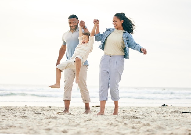 Rodzina na plaży i trzymanie się za ręce na świeżym powietrzu, aby zapewnić sobie swobodę podróżowania i więź w naturze. Kochaj zabawę i chłopca z młodymi rodzicami nad morzem, grając w huśtawkę i szczęśliwych na wakacjach nad oceanem