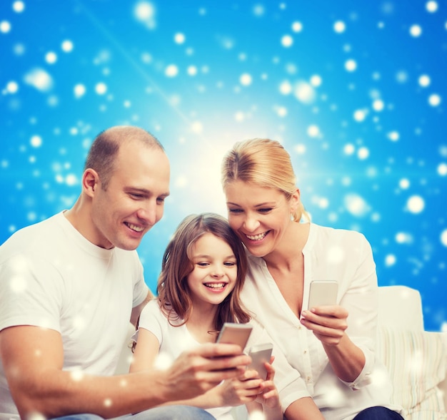 rodzina, święta Bożego Narodzenia, technologia i koncepcja ludzi - uśmiechnięta matka, ojciec i mała dziewczynka ze smartfonami na niebieskim śnieżnym tle