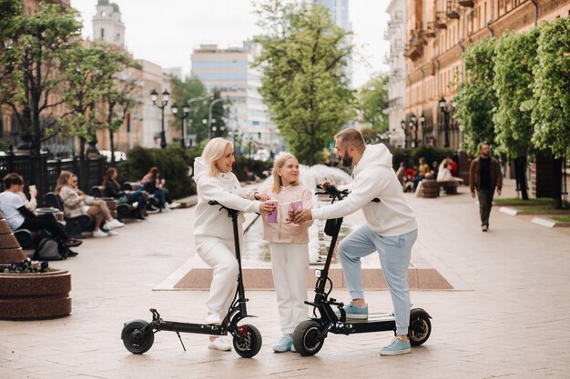 Rodzina w białych ubraniach stoi w mieście na elektrycznych hulajnogach i pije drinka.