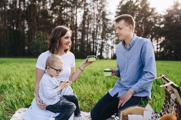 Rodzina W Lesie Na Pikniku. Usiądź Na Polanie, Zielonej Trawie. Niebieskie Ubrania. Mama I Tata Bawią Się Z Synem, Obejmują Się I Uśmiechają.