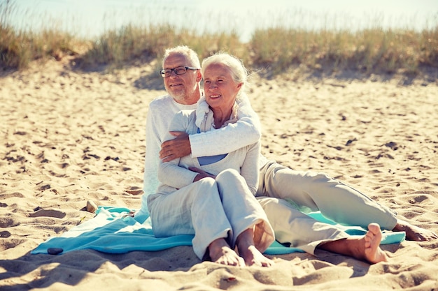 rodzina, wiek, podróże, turystyka i ludzie koncepcja - szczęśliwa para seniorów siedząca na placu i uściskająca się na letniej plaży