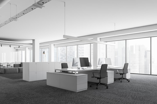 Róg białego biura z panoramicznymi oknami, szary dywan na podłodze, szare stoły komputerowe z czarnymi krzesłami i szafkami.