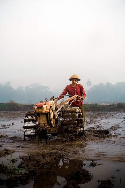 Rolnik w czerwonej koszuli i czerwonym kapeluszu stoi na polu z traktorem, który mówi „rolnik pracuje”.