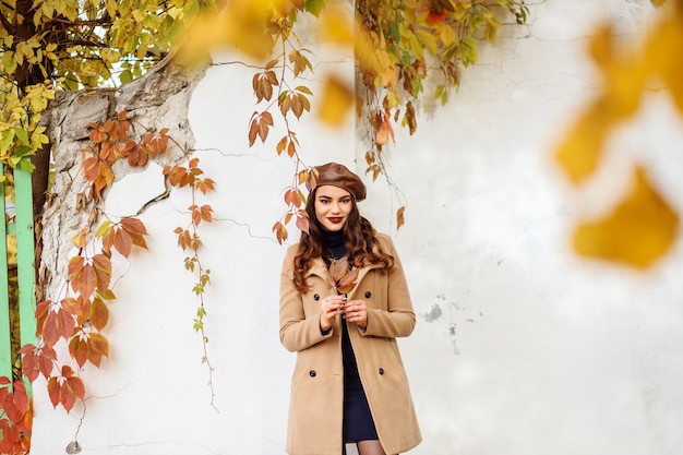 Romantyczna kaukaska kobieta, ubrana w brązowy beret i beżowy płaszcz, jesienią spędza czas na świeżym powietrzu. Z przodu żółte, niewyraźne liście.