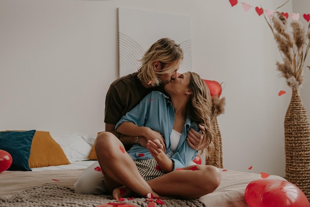 Romantyczna kochająca para siedząca na łóżku i całująca się, podczas gdy czerwone konfetti w kształcie serca latają