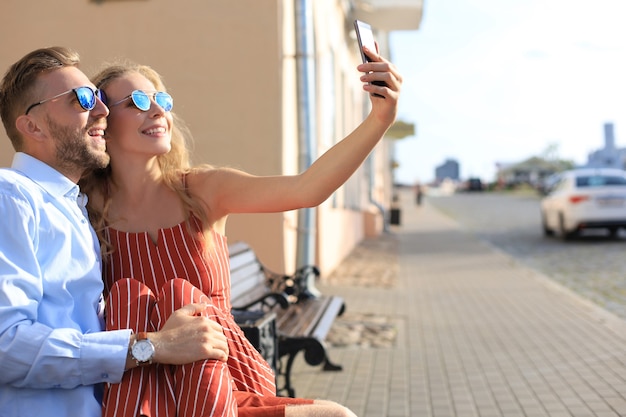 Romantyczna młoda para w letnie ubrania, uśmiechając się i biorąc selfie siedząc na ławce na ulicy miasta.