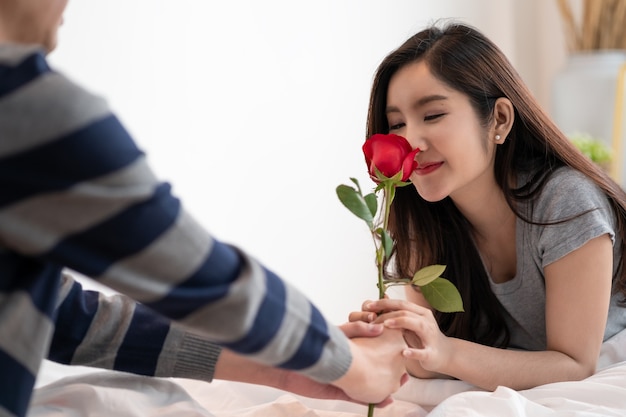 Romantyczna Para Azjatycka W Sypialni, Mężczyzna Dający Różę Pięknej Kobiecie I Oboje Całujący Piękną Różę Z Miłością I Szczęściem, Piękna Elegancka Para Azjatycka Przytula Się I Uśmiecha Się W Sypialni