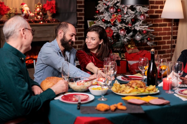 Romantyczna para świętująca Boże Narodzenie z rodziną, szczęśliwym chłopakiem i dziewczyną rozmawiającą przy świątecznym stole. Święta Bożego Narodzenia, żona i mąż jedzą tradycyjny posiłek z rodzicami