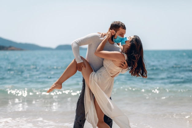Romantyczna para żartobliwie całuje się na plaży w medycznych maskach, które chronią ich przed wybuchem infekcji koronawirusem. Mężczyzna trzymający dziewczynę za nogę i całujący ją
