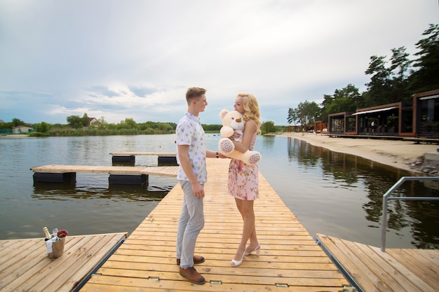 Romantyczna randka niespodzianka. Młody chłopak i dziewczyna na molo z widokiem na jezioro. Facet daje dziewczynie misia.