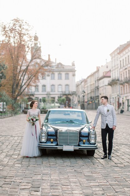 Romantyczna ślub pary pozycja przed czarnym retro samochodem, piękny szczęśliwy państwo młodzi w lecie, starzy miasto budynki na tle