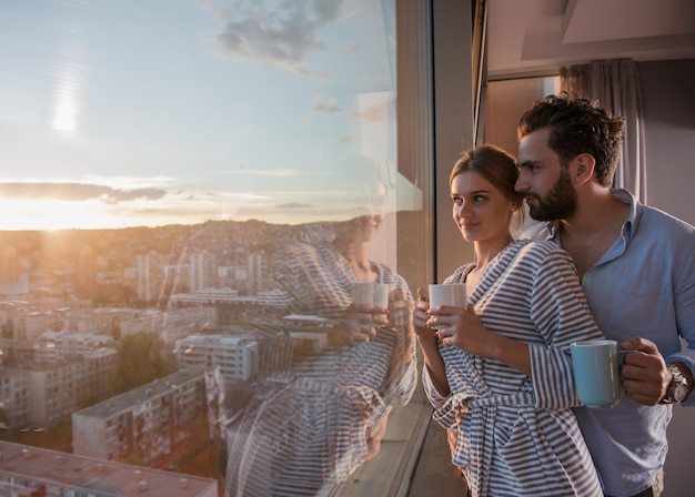romantyczna szczęśliwa młoda para ciesząca się wieczorną kawą i pięknym zachodem słońca krajobraz miasta, stojąc przy oknie