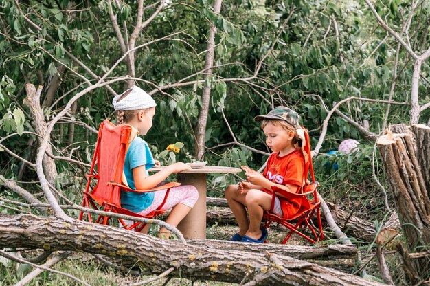 Rosja, Lipiec 2020 - Małe Dzieci, Chłopiec I Przyjaciółka Urządzają Piknik I Jedzą Wśród Powalonych Drzew Latem Na Wiosce Na Wsi Na Wakacjach