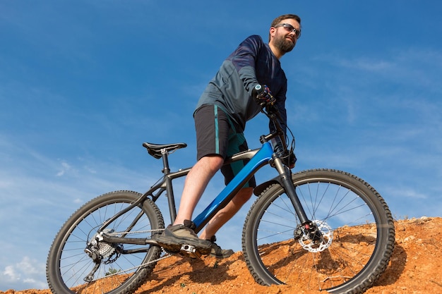 Rowerzysta W Krótkich Spodenkach I Koszulce Na Nowoczesnym Karbonowym Rowerze Hardtail Z Widelcem Pneumatycznym Stojącym Na Klifie Na Tle świeżego Zielonego Letniego Lasu