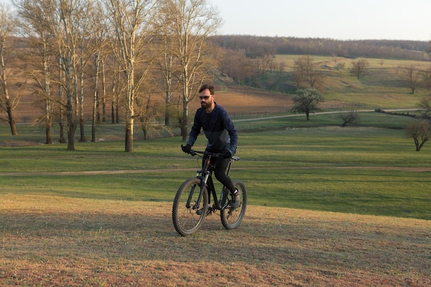 Rowerzysta w krótkich spodenkach i koszulce na nowoczesnym karbonowym rowerze hardtail z widelcem pneumatycznym stojącym na klifie na tle świeżego zielonego wiosennego lasu