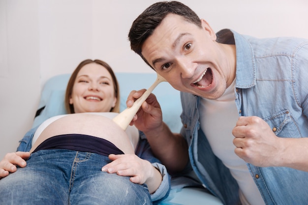 Rozbawiony szczęśliwy, zachwycony mąż siedzi w klinice i używa sprzętu medycznego, słuchając bicia serca płodu w brzuszku w ciąży