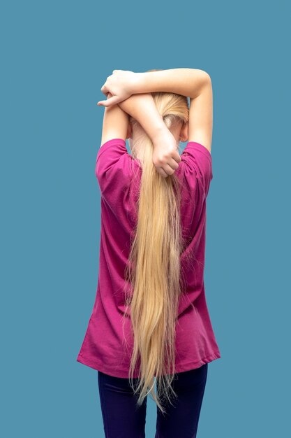 Rozciąganie, sport. Widok z tyłu dziewczyny z długimi blond włosami w odzieży sportowej robi rozciąganie ramion na niebieskim tle