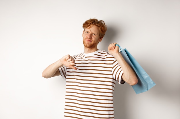 Rozczarowany młody mężczyzna z rudymi włosami i brodą pokazujący kciuk w dół po złych zakupach, trzymający torbę przez ramię i marszczący brwi, zdenerwowany, białe tło