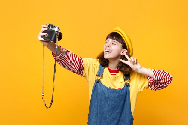 Roześmiana dziewczyna nastolatka we francuskim berecie z zamkniętymi oczami robi selfie nakręcony na retro vintage aparat fotograficzny, pokazując znak zwycięstwa na białym tle na żółtym tle. Ludzie szczere emocje, koncepcja stylu życia.